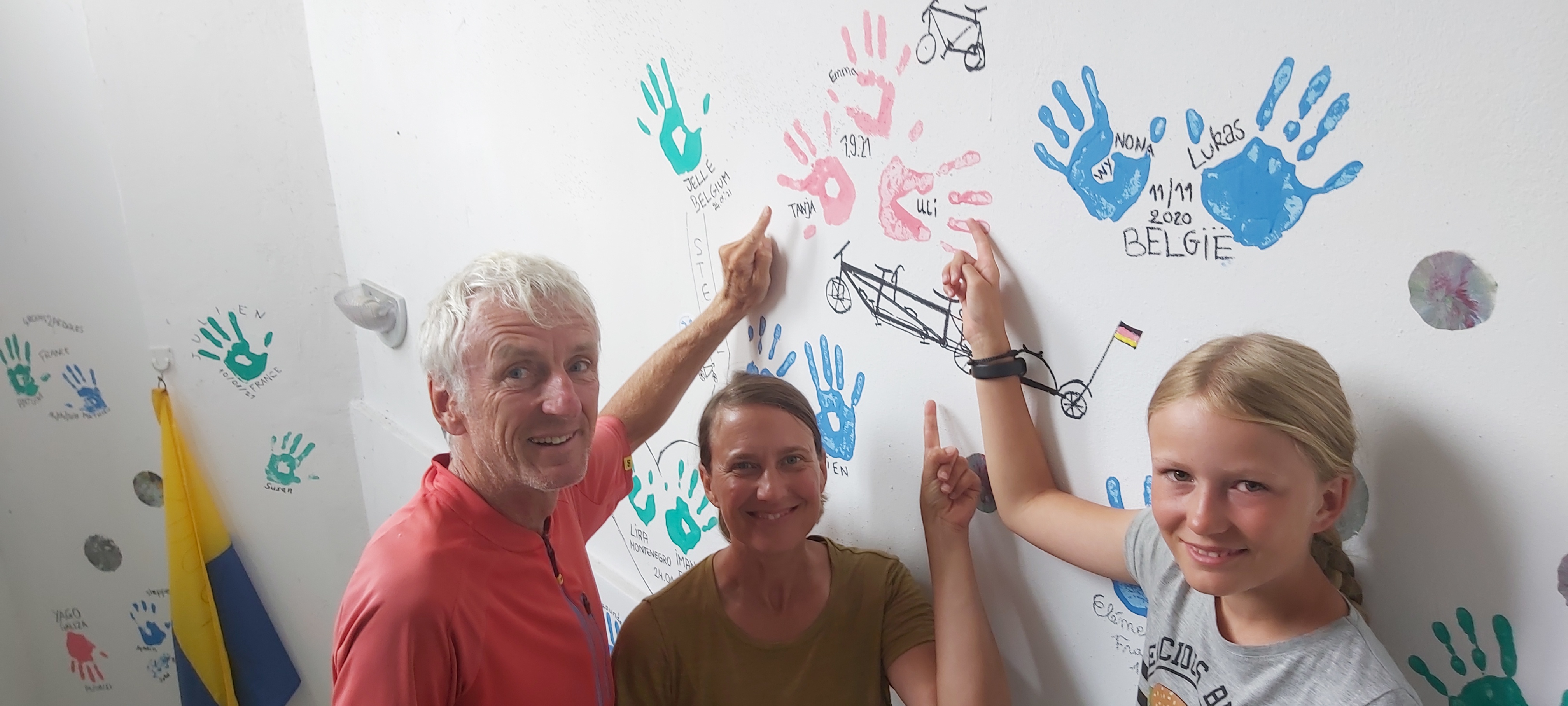 Drei Personen zeigen auf eine Wand mit aufgemalten Handabdrücken vergangener Gäste.